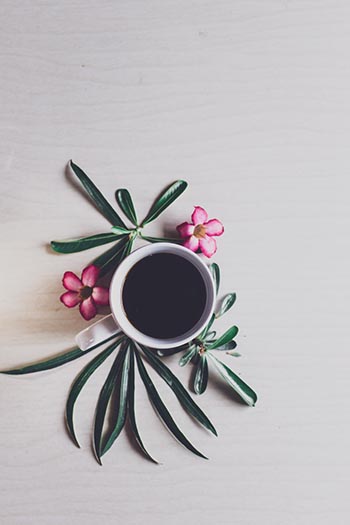 A cup of black coffee rests on a surface surrounded by tropical pink and green flowers.